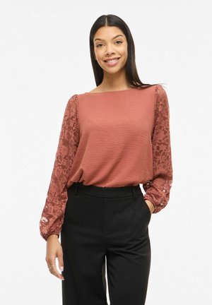 Nainen hymyillen, yllä ruosteenvärinen teksturoidusta kankaasta valmistettu pusero, jossa pitsiset pitkät hihat, ja mustat korkeavyötäröiset housut, seisomassa käsi taskussa.