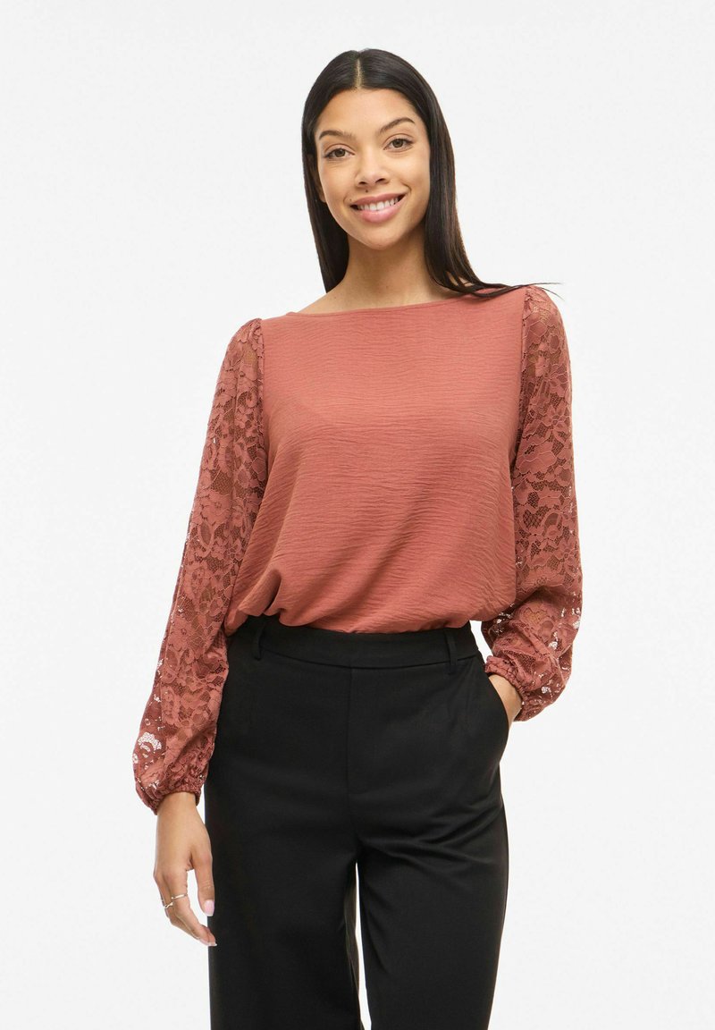 Femme souriante portant un chemisier texturé couleur rouille avec des manches longues en dentelle et un pantalon noir taille haute, debout avec une main dans la poche.