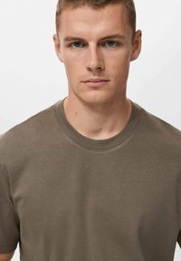 Ung mand med kort lyst hår og blå øjne iført en ensfarvet olivengrøn t-shirt med rund hals, der står vendt fremad mod en hvid baggrund.