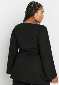 Top nero strutturato con maniche lunghe e svasate e vita con cintura. Presenta un semplice scollo rotondo e un tessuto con un leggero motivo.