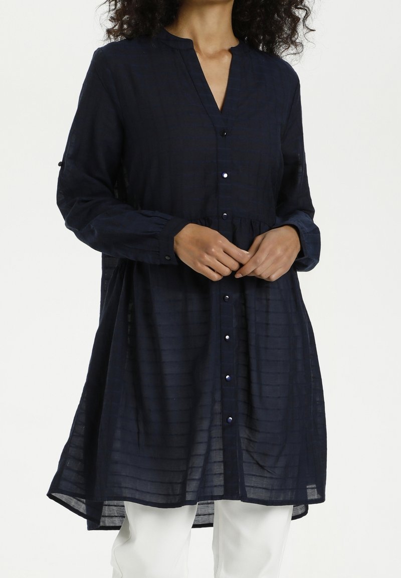 Femme portant une tunique bleu marine foncé à manches longues, boutonnée, avec des rayures horizontales subtiles, associée à un pantalon blanc, sur un fond uni.