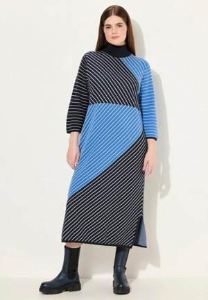 Lange, gekleurde jurk met kleurblokken en diagonale strepen in blauw en zwart, met een hoge hals en driekwart mouwen, gecombineerd met zwarte enkellaarsjes.