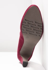 Chaussure à talon haut en daim rouge présentée semelle vers le haut, affichant la marque "Tamaris" et la taille 37 sur une semelle marron lisse.