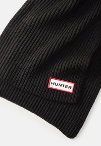 Černý žebrovaný pletený šátek s červenou a bílou štítkem s nápisem "HUNTER". Měkká textura a prodloušený obdélníkový tvar.