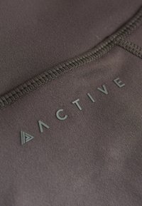Tamsiai pilkos spalvos aktyviosios aprangos audinys, pasižymintis plokščiomis siūlėmis ir pakeltu plastiku užrašytu žodžiu "ACTIVE" su trikampio formos logotipo akcentu.