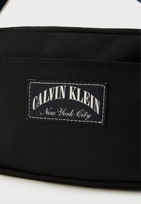 Μαύρη υφασμάτινη τσάντα με ορθογώνια ετικέτα που φέρει το λογότυπο "CALVIN KLEIN" με έντονα λευκά γράμματα και "New York City" σε μικρότερα γράμματα, περιβαλλόμενη από λευκό ραφτό περίγραμμα.