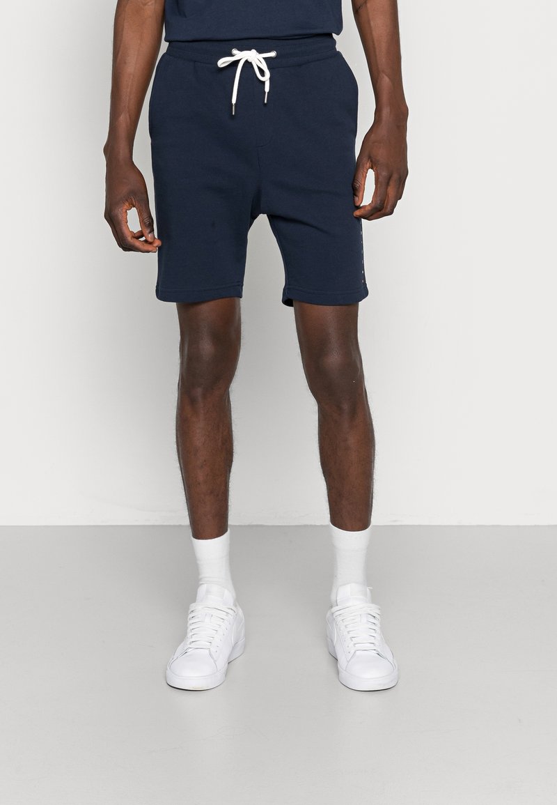 Shorts en mélange de coton bleu marine avec une taille élastique et un cordon de serrage blanc. Dotés de poches latérales et d'une coupe slim. Associés à des baskets blanches et des chaussettes.