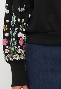 Manche d'un pull noir avec broderie florale colorée à côté d'un jean en denim bleu, mettant l'accent sur la zone du poignet et de la taille.