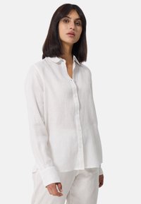 Camicia bianca di lino con design a bottoni, maniche lunghe e vestibilità rilassata. Tessuto texture con un aspetto morbido e traspirante.
