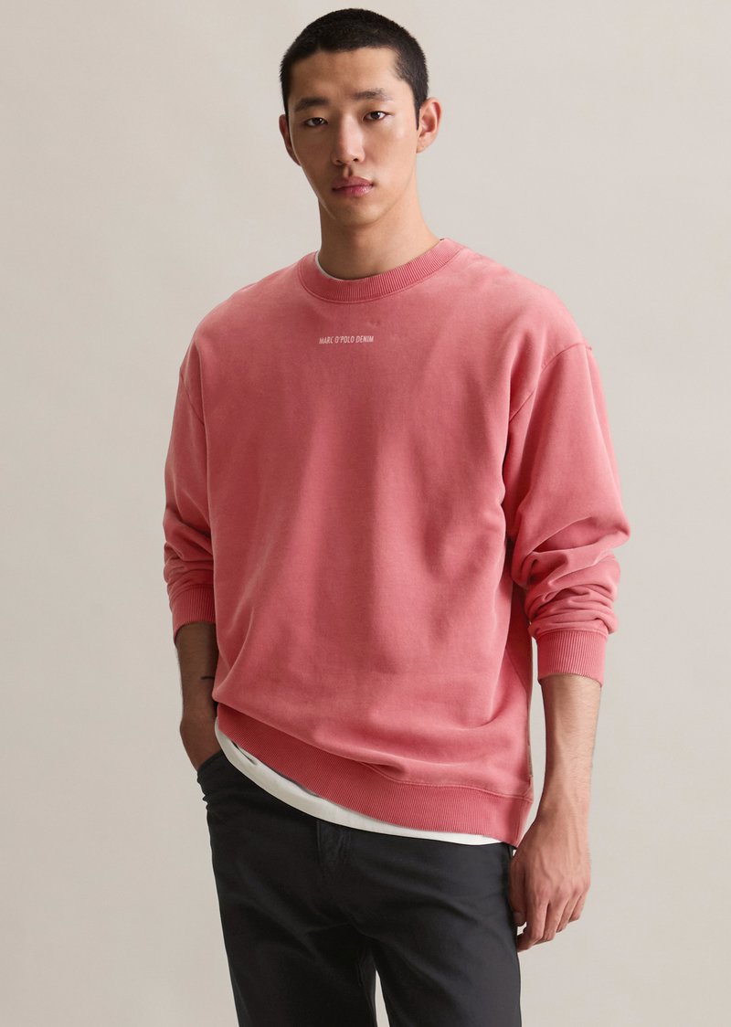 Sweatshirt rose à col rond en tissu doux, présentant des poignets et un ourlet côtelés, avec un logo sur la poitrine et porté par-dessus une chemise blanche.