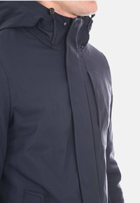 Giacca impermeabile blu navy con colletto alto, cappuccio regolabile e chiusura frontale con zip. Presenta una tasca sul petto e una texture del tessuto liscia.