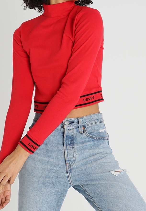 Haut court rouge à manches longues avec un col montant et bande noire avec le logo "Levi's" à l'ourlet et aux poignets, associé à un jeans en denim déchiré bleu clair.