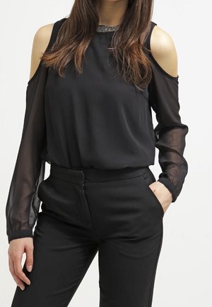 Femme portant un chemisier noir à épaules dénudées avec manches longues transparentes et un pantalon noir taille haute, mains dans les poches.