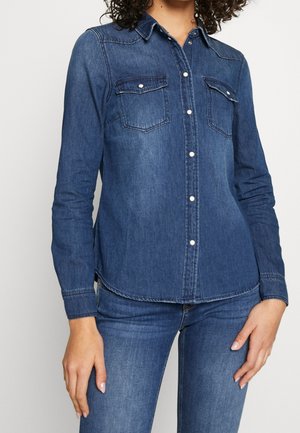 Frau trägt ein eng anliegendes blaues Jeanshemd mit Druckknöpfen und zwei Klappentaschen auf der Brust, kombiniert mit blauen Jeans.