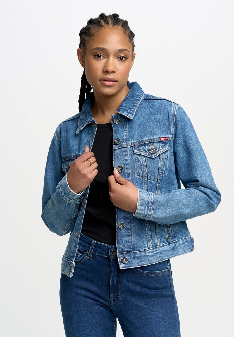 Veste en denim bleu clair avec fermeture à boutons, poches poitrine et coupe décontractée. Portée sur un haut noir et associée à un jean bleu.