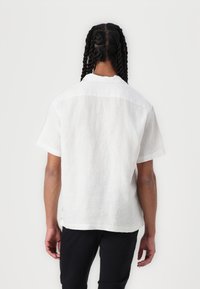 Chemise blanche en lin à manches courtes avec une coupe droite et une texture subtile. Présente un col classique et un détail de dos.
