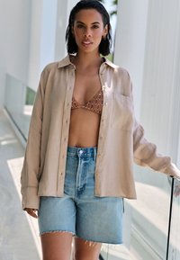 Light beige button-up shirt made of soft fabric, worn over a patterned brown bikini top and paired with frayed blue denim shorts.