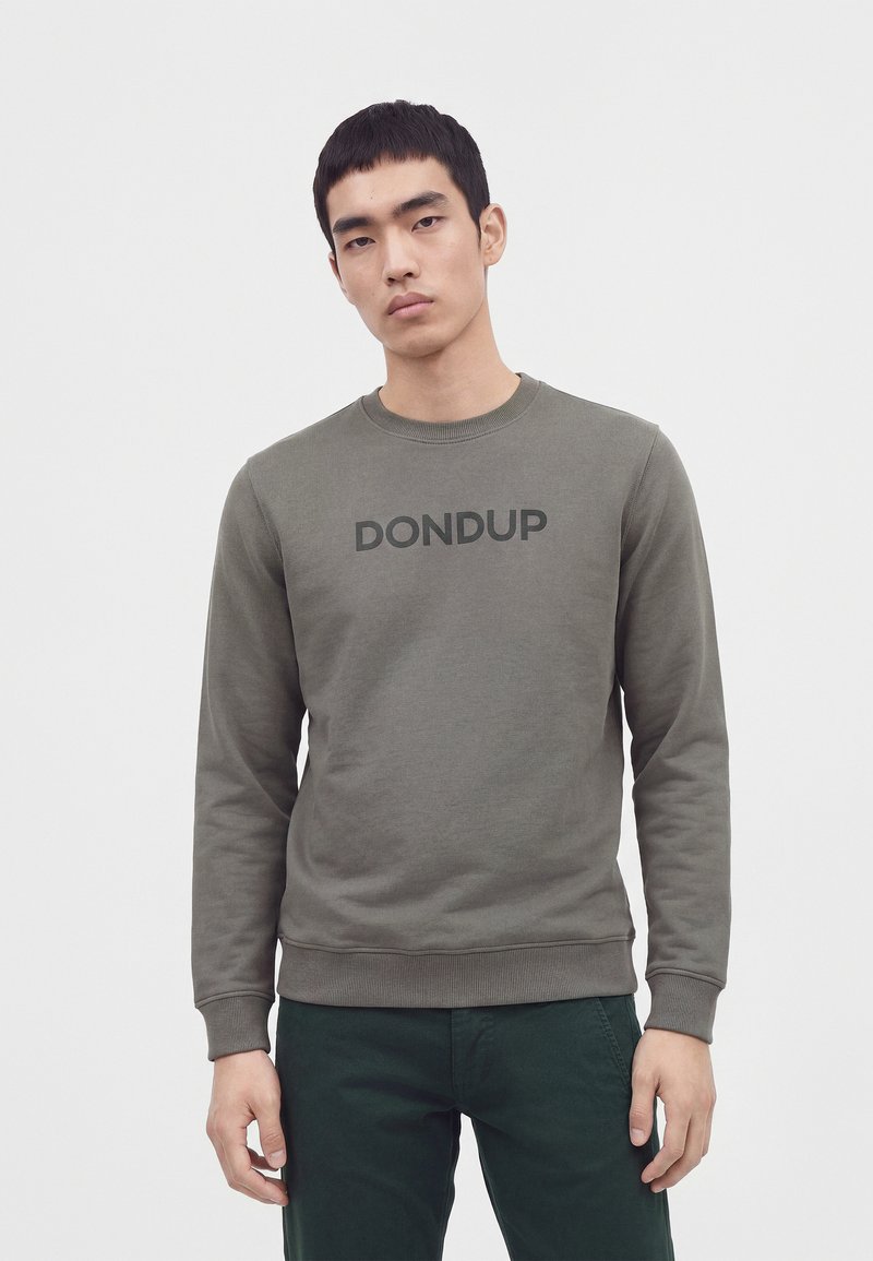 Jeune homme portant un sweat-shirt gris avec le texte "DONDUP" et un pantalon vert foncé, debout devant un fond blanc uni.