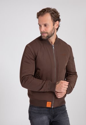 Man met baard en oorbel die een bruine bomberjack met rits en donkere spijkerbroek draagt, staat tegen een effen lichte achtergrond.