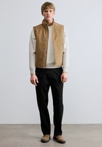 Rjavkasta antilop brezrokavnik z zadrgo spredaj in visokim ovratnikom čez svetlo sivi rebrasti pulover. Črne hlače dokončajo obleko, z bež čevlji. Minimalistični slog.