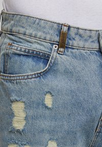 Jeans din denim ușor albastru, uzate, cu petice destrămate, un nasture din alamă și un design clasic cu cinci buzunare. Textura pare uzată și moale.
