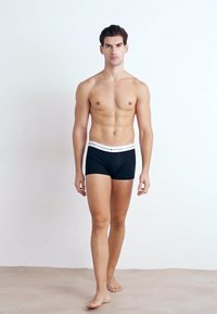 Schwarze Baumwoll-Boxershorts mit einem weißen Bund, eng geschnittenes Design und glatter Textur; Modell steht barfuß vor einem hellen Hintergrund.