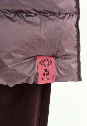 Textilní štítek na levandulově fialové polstrované bundě s grafikou oka a nápisem "SEE IT ALL NAOKO" na bílém pozadí.