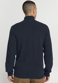 Marinblå stickad tröja med hög krage, texturerat mönster på axlar och ärmar samt ribbade muddar och nederkant.