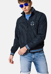 Granatowa nylonowa kurtka bomberka z zapięciem na zamek, białym haftem logo i paskowanymi mankietami. Połączona z jasnoniebieskimi szortami dżinsowymi.