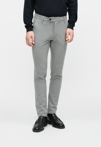 Homme portant un pantalon slim gris clair, un top noir à manches longues et des chaussures en cuir noires, debout devant un fond uni.