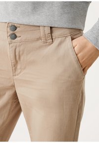 Pantalon beige avec une texture lisse, doté de deux boutons gris à la taille, de poches latérales et d'une coupe ajustée.