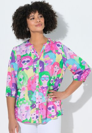 Femme aux cheveux bouclés portant une chemise colorée à boutons ornée de visages abstraits avec des lunettes de soleil et des motifs floraux, assortie à un pantalon blanc.