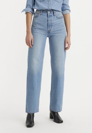 Femme portant un jean taille haute bleu clair à jambes droites, une chemise boutonnée bleu clair aux manches retroussées, et des bottines noires à talons.