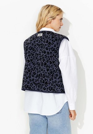 Gilet en imprimé léopard bleu marine avec une surface texturée, présentant une étiquette au dos et porté sur une chemise blanche, associé à un jean bleu.