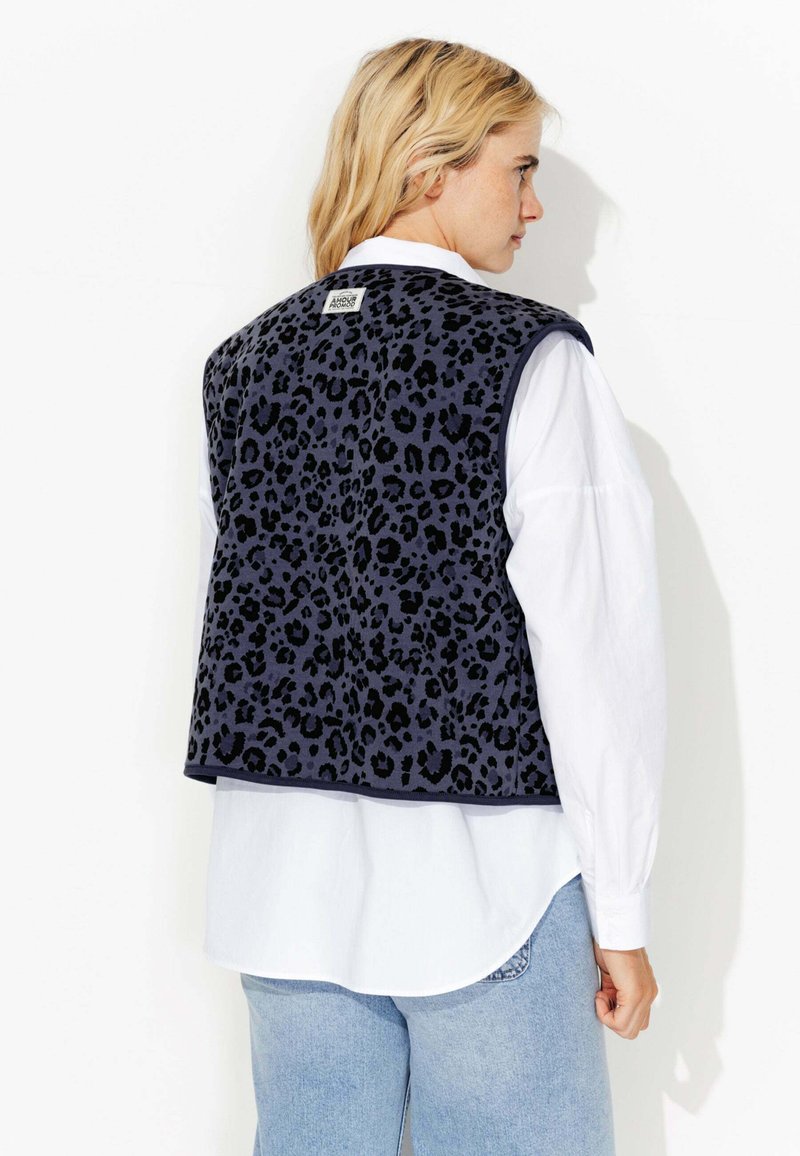 Gilet en imprimé léopard bleu marine avec une surface texturée, présentant une étiquette au dos et porté sur une chemise blanche, associé à un jean bleu.