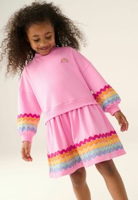 Rosa Sweatshirt-Kleid mit langen Ärmeln, ausgestattet mit einem Regenbogen-Patch und welligem, mehrfarbigem Saum an den Ärmeln und am unteren Rand. Weicher Stoff.