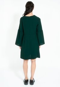 Zelena haljina do koljena s oblikovanim rukavima, zaobljenim izrezom i zatvaračem na leđima. Glatka tkanina s suptilnim strujanjem i pokretom.