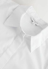 Biela košeľa s golierom, hladká na dotyk, v klasickom strihu. Na límci je viditeľná visačka s informáciami o značke a starostlivosti.