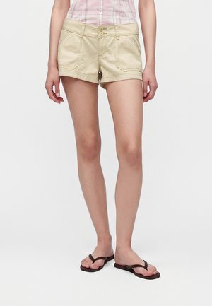 Femme portant un short beige, une chemise à carreaux rose et des tongs marron foncé, se tenant devant un fond blanc uni.