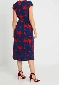 Kvinna iklädd en marinblå midiklänning med röd blommig design och vinröda högklackade ankelremsskor, stående mot en enfärgad bakgrund.