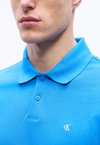Νεαρός άνδρας που φοράει φωτεινό μπλε πόλο πουκάμισο Calvin Klein με γιακά και δύο κουμπιά κλειστά, το λογότυπο φαίνεται στο στήθος.