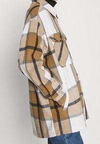 Camicia a quadri in beige, bianco e grigio. Presenta un colletto, taschini sul petto e chiusura frontale con bottoni. Realizzata in materiale flanella con una texture morbida.