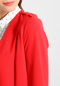 Haut léger en tissu rouge avec manches amples, comprenant un détail de bouton sur l'épaule et un col avec des pois noirs en dessous.