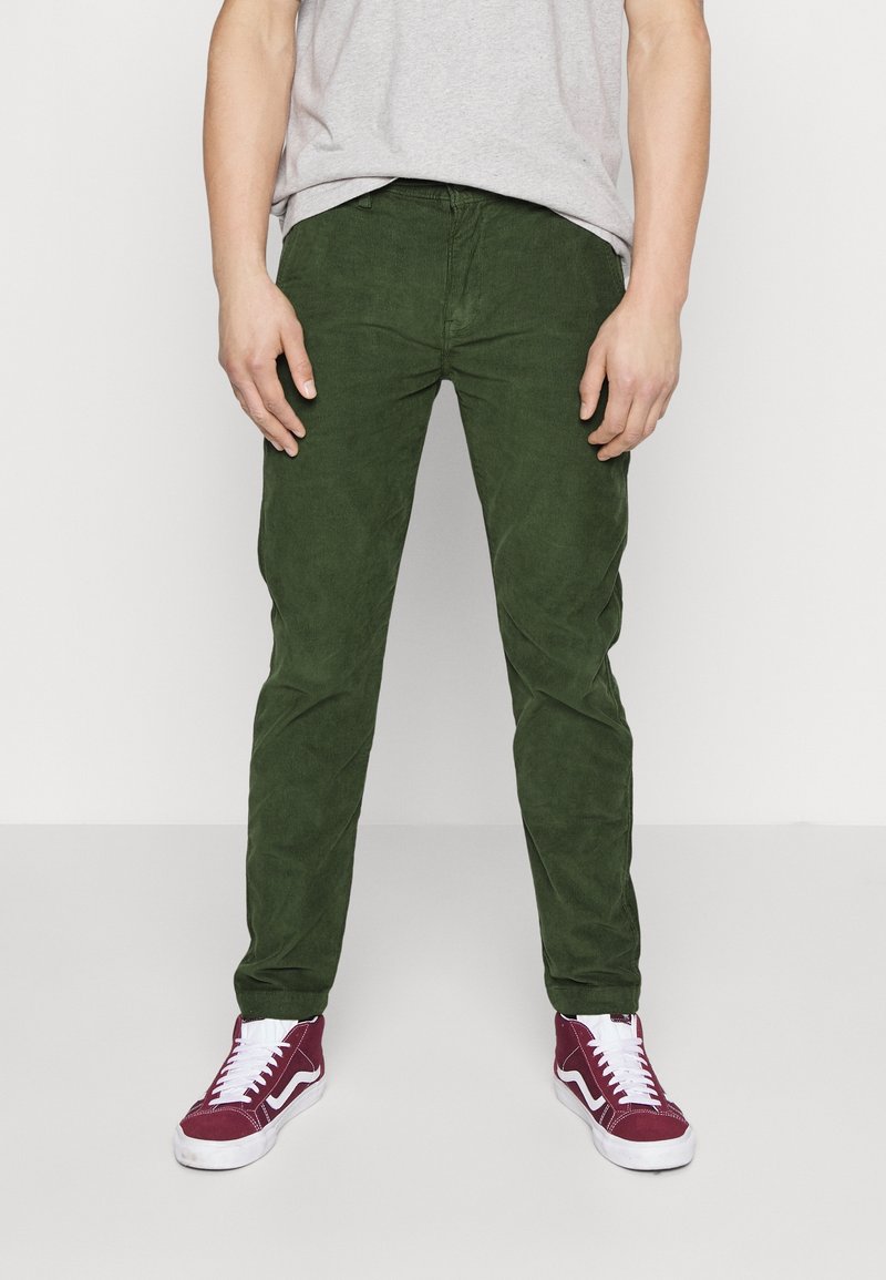 Homme portant un pantalon en velours côtelé vert, chemise grise et baskets montantes bordeaux, debout devant un fond blanc uni.
