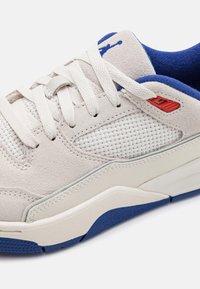 Sneaker bianca in suede con pannelli in rete, dettagli blu e rossi. Presenta lacci piatti e una suola in gomma strutturata per una maggiore aderenza.