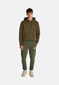 Sudadera verde oliva con un bolsillo frontal y parche de logo, combinada con joggers oliva que presentan un logo lateral, usados con zapatillas beige.