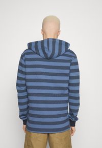Randig blå hoodie med växlande mörka och ljusa ränder, texturerad tygkvalitet och dragsko i huvan. Långa ärmar med ribbad manschett.
