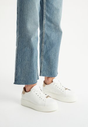 Sneaker bianchi in pelle con finitura testurizzata, dotati di dettagli in oro e una suola spessa, indossati con jeans in denim blu chiaro e cropped.