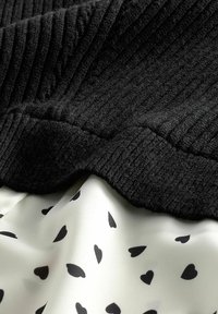 Tessuto a coste nero sovrapposto a un tessuto color crema con piccoli motivi a forma di cuore neri. Le texture sono distinte e contrastanti.