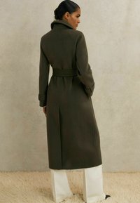 Oliivroheline villane mantel kõrge kraega, vöödiga talje ja pikkade varrukatega; omab sirget lõiget ja sujuvat tekstuuri.